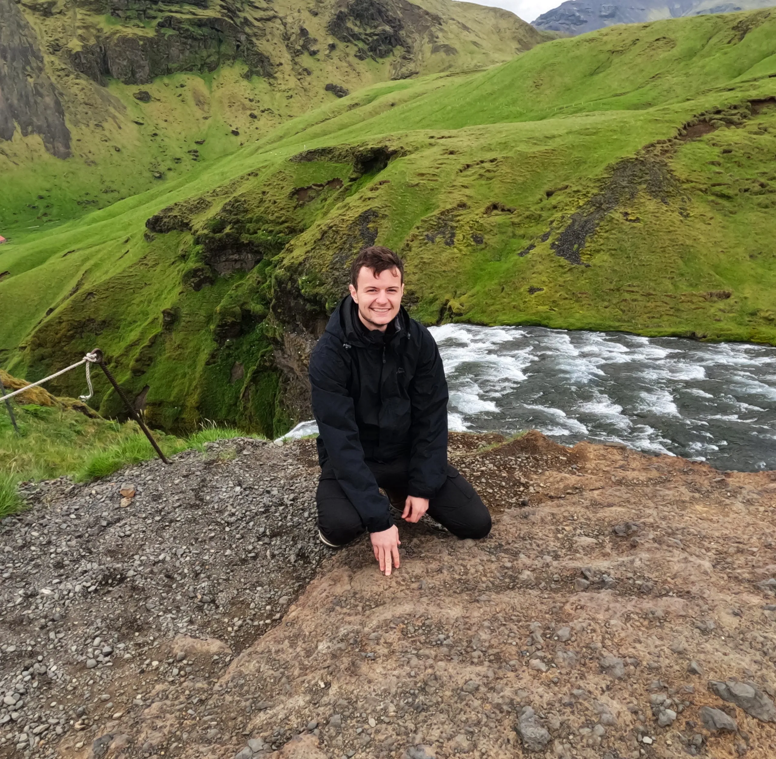 Jacob smiling as he is crouched down on a rocky path right next to a waterfall. Behind him there is the top of the waterfall and green mountains.