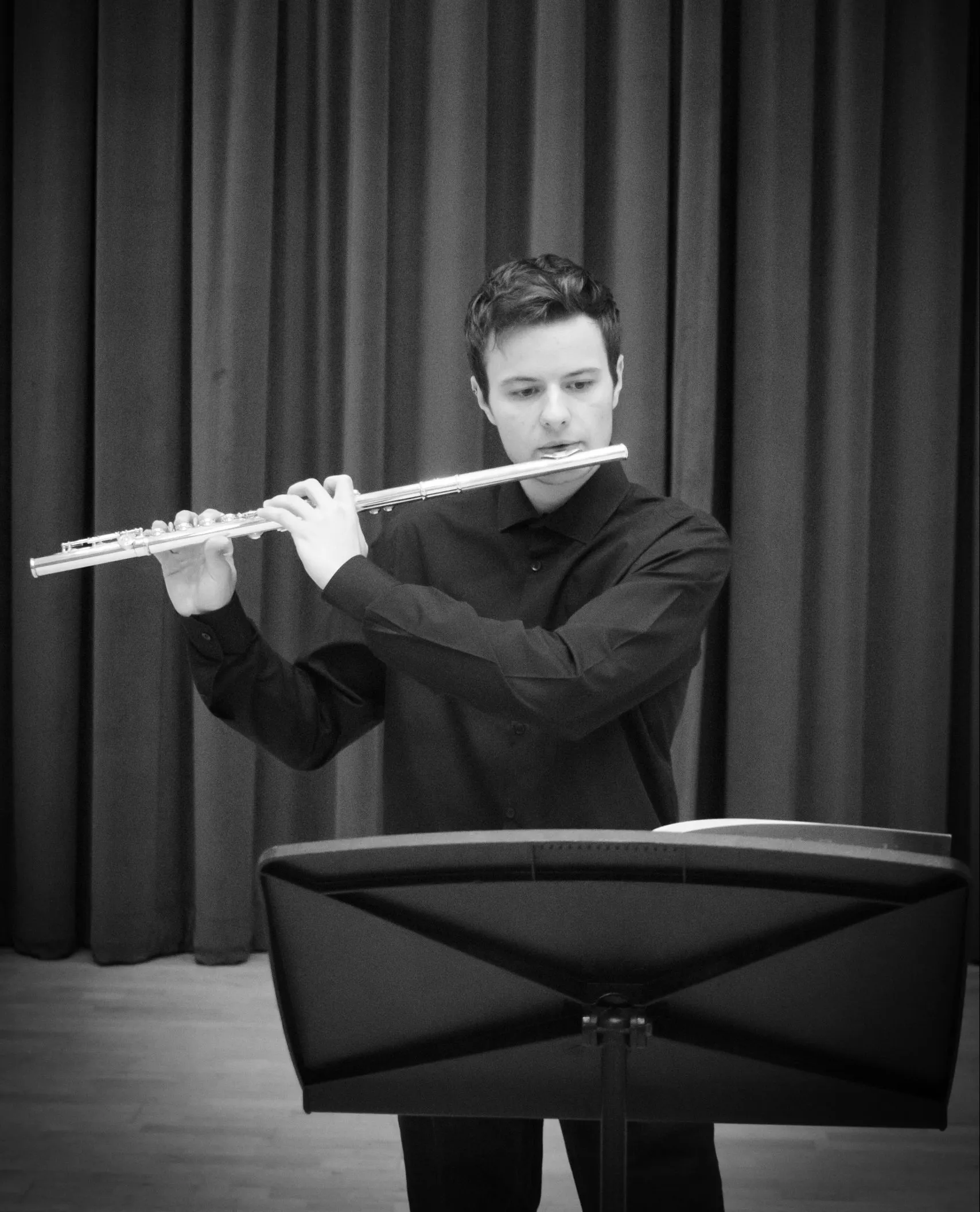 Black & White image of Jacob playing flute. He is standing on a stage with a big curtain behind him and a music stand in front of him.