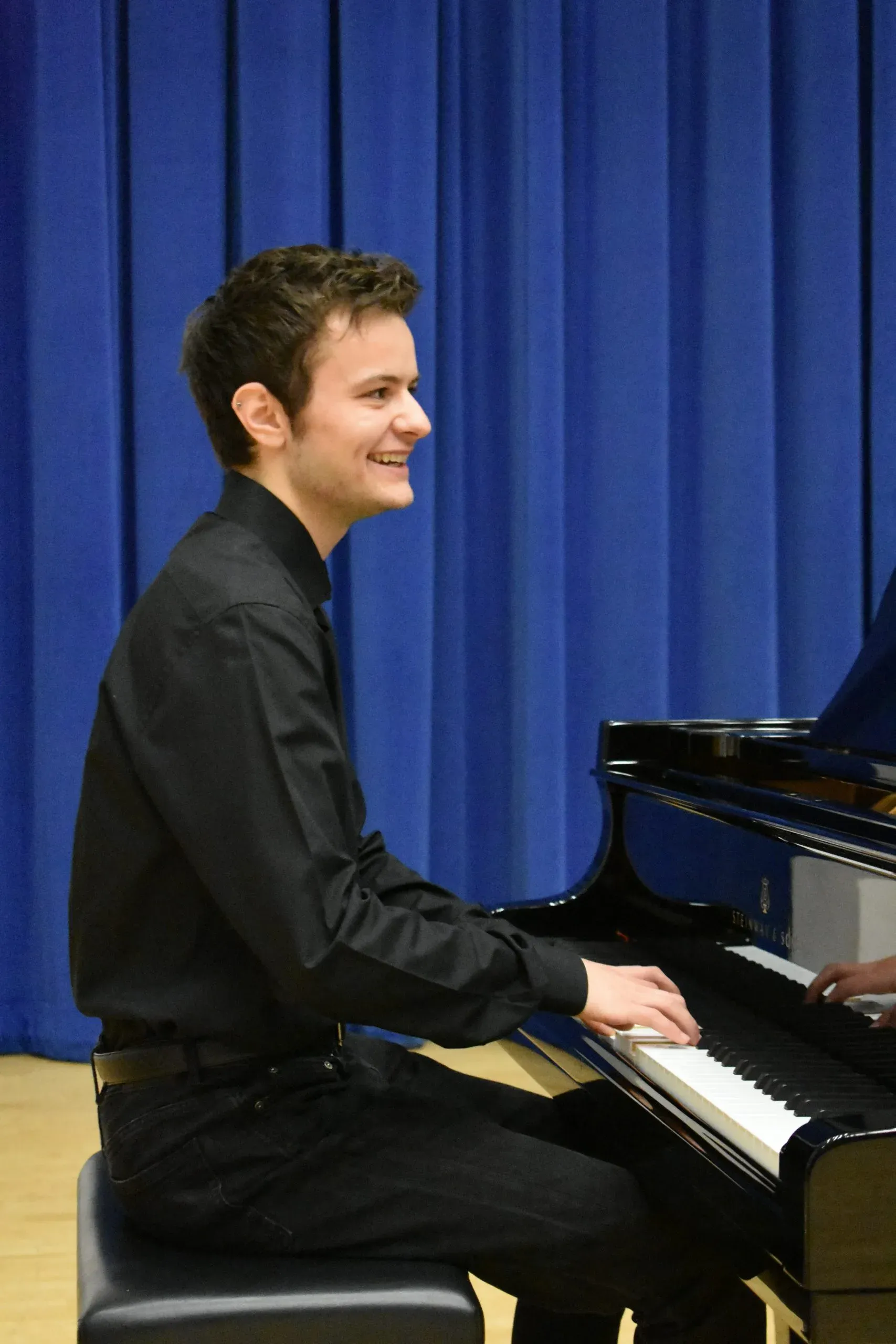 Jacob sitting at a Steinway grand piano, wearing black shirt and trousers, with a blue curtain behind him. He is playing on stage and smiling at someone off camera