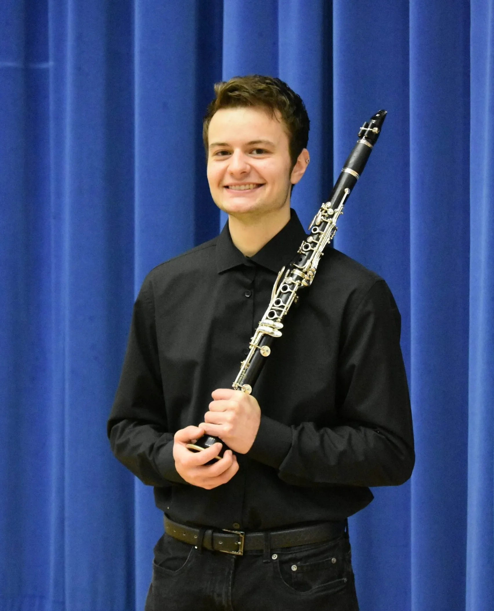 Jacob standing in front of a blue curtain wearing a black shit and trousers. He is holding his clarinet and smiling.