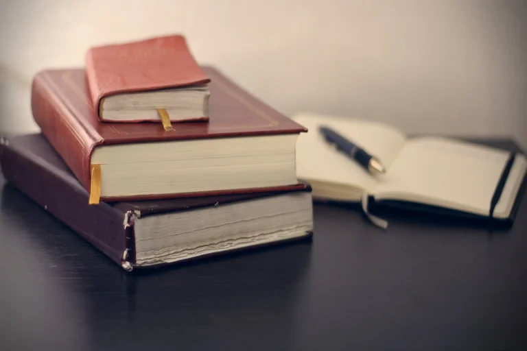 Stack of 3 books of varying shades of brown with an open diary in the background with a pen sitting on top of it.