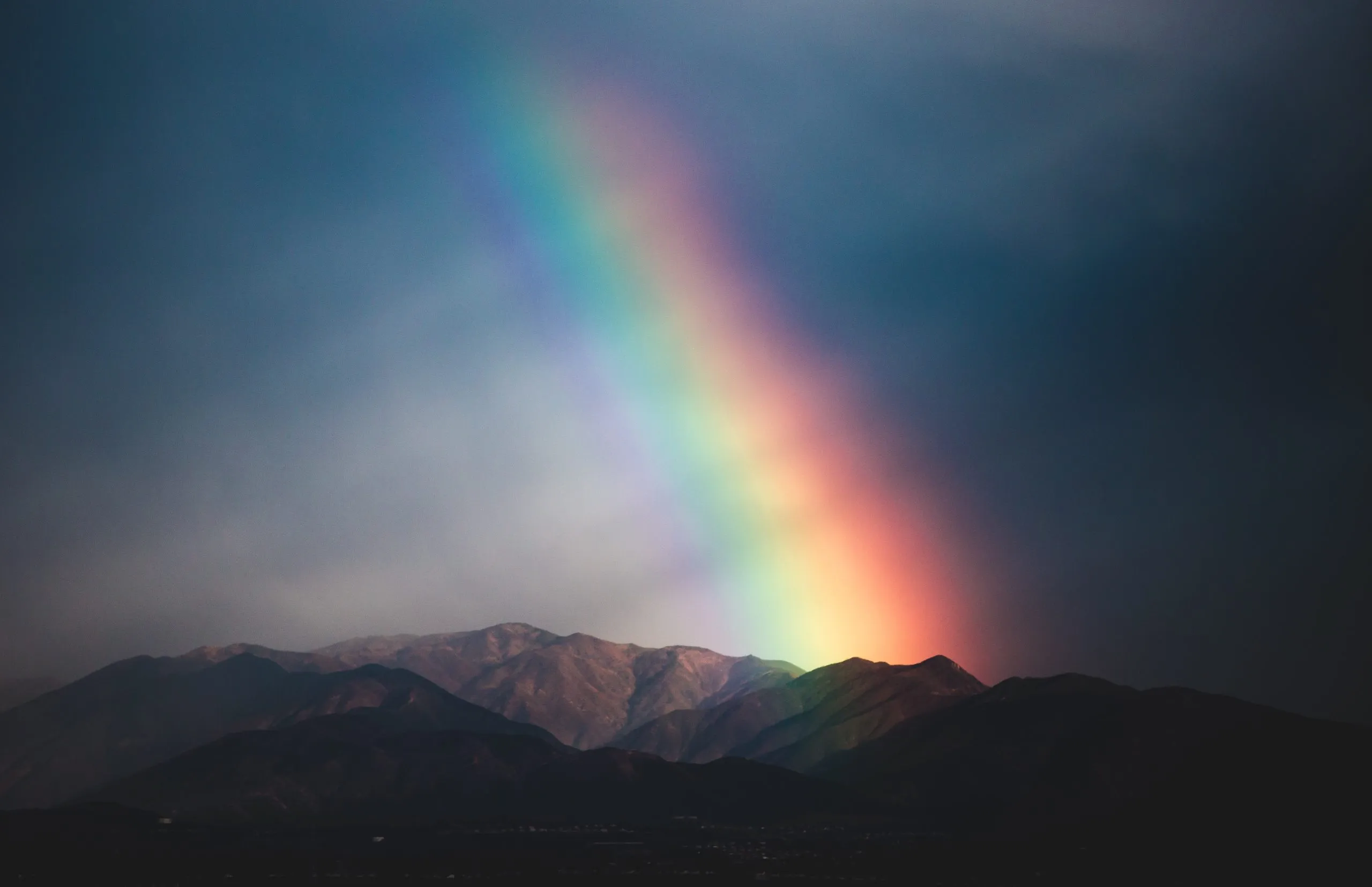 A dark image of mountains with a dark sky. Above the mountains in the middle of the image there is a rainbow.